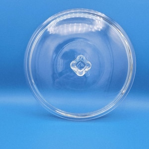 Może przedstawiać: Przezroczysta szklana pokrywka o zaokrąglonym kształcie z małym, czteropłatkowym uchwytem w kształcie kwiatu pośrodku. Pokrywka jest przeznaczona do okrągłego pojemnika. Tło jest jednolicie niebieskie.