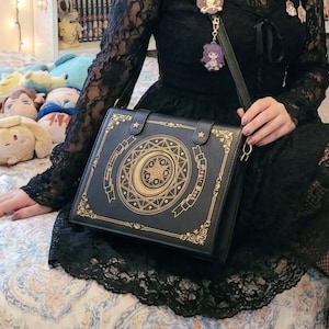 May include: Black shoulder bag with a book-like design. The bag features gold embellishments, including a crescent moon and star pattern, and the words "Magic Book". The bag has a long strap and a small charm.