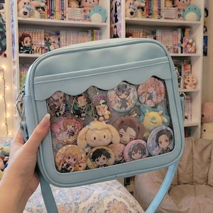 May include: A light blue crossbody bag with a clear front display area, filled with character badges and plushies. The bag has a scalloped edge design and a long shoulder strap. The bag is filled with various anime-style character badges and small plush toys.