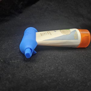 Puede incluir: Un exprimidor de tubos de plástico azul con un tubo de producto blanco y naranja. El tubo tiene una etiqueta con texto y un gráfico. El exprimidor está diseñado para ayudar a dispensar el producto.