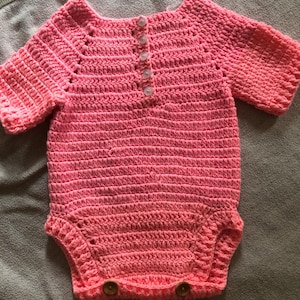 Puede incluir: Un pelele de ganchillo rosa brillante con mangas cortas, cuello redondo y una fila vertical de cuatro botones transparentes pequeños en la parte superior del pecho. Las aberturas de las piernas tienen dos botones de madera marrones.