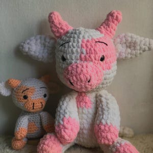 Può includere: Due peluche di mucca all'uncinetto fatti a mano. La mucca più grande è bianca e rosa, con orecchie, zoccoli e muso rosa. La mucca più piccola è grigia e pesca. Entrambe hanno occhi neri e sono sedute su una superficie bianca e morbida.