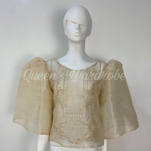 Puede incluir: Una blusa color crema con mangas anchas y transparentes y bordados intrincados en el corpiño. La blusa se exhibe en un maniquí sobre un fondo blanco. El texto "Queen's Wardrobe" es visible en la blusa.