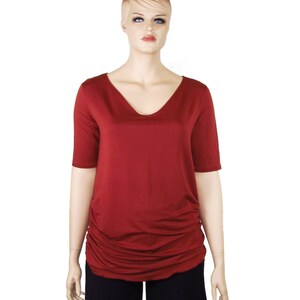 May include: A maroon V-neck top with short sleeves and a gathered side detail. The top is displayed on a mannequin, paired with black pants. The fabric appears soft and drapes well, suggesting a comfortable fit. The overall style is casual and versatile.