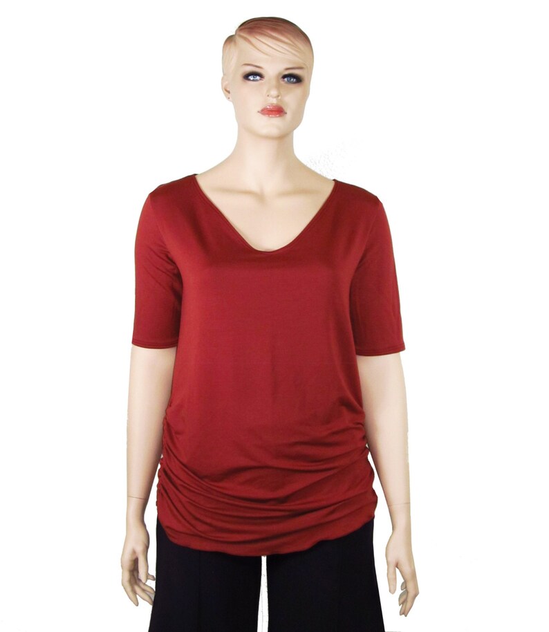 May include: A maroon V-neck top with short sleeves and a gathered side detail. The top is displayed on a mannequin, paired with black pants. The fabric appears soft and drapes well, suggesting a comfortable fit. The overall style is casual and versatile.