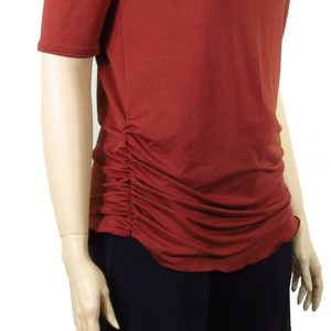 May include: Rust-colored short-sleeved top with a ruched side detail. The top has a scoop neckline and a slightly curved hemline. The fabric appears to be a soft, lightweight material. The top is paired with black pants.