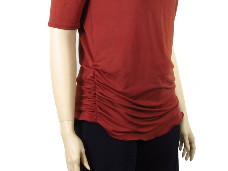 May include: Rust-colored short-sleeved top with a ruched side detail. The top has a scoop neckline and a slightly curved hemline. The fabric appears to be a soft, lightweight material. The top is paired with black pants.