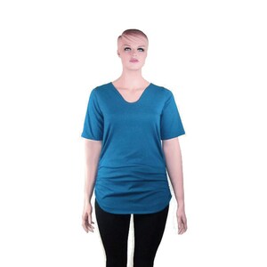 May include: A teal V-neck short-sleeve shirt with a gathered side detail. The shirt has a curved hemline and is displayed on a mannequin. The mannequin is wearing black pants.