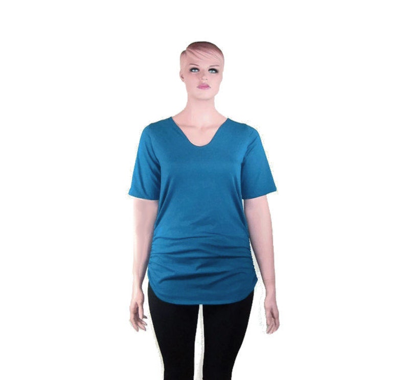 May include: A teal V-neck short-sleeve shirt with a gathered side detail. The shirt has a curved hemline and is displayed on a mannequin. The mannequin is wearing black pants.