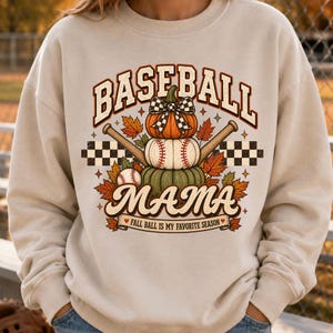 May include: A beige crewneck sweatshirt featuring a graphic print of three stacked pumpkins, two crossed baseball bats, a baseball, autumn leaves, and a checkered pattern. The text reads BASEBALL MAMA and FALL BALL IS MY FAVORITE SEASON.