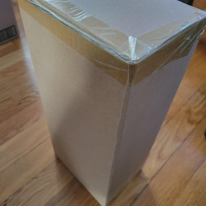 Puede incluir: Una caja de cart&oacute;n rectangular y alta, sellada con cinta adhesiva transparente. La caja es de color marr&oacute;n claro con una parte superior de color m&aacute;s claro. Una peque&ntilde;a etiqueta es visible en la parte superior. La caja est&aacute; sobre un suelo de madera.