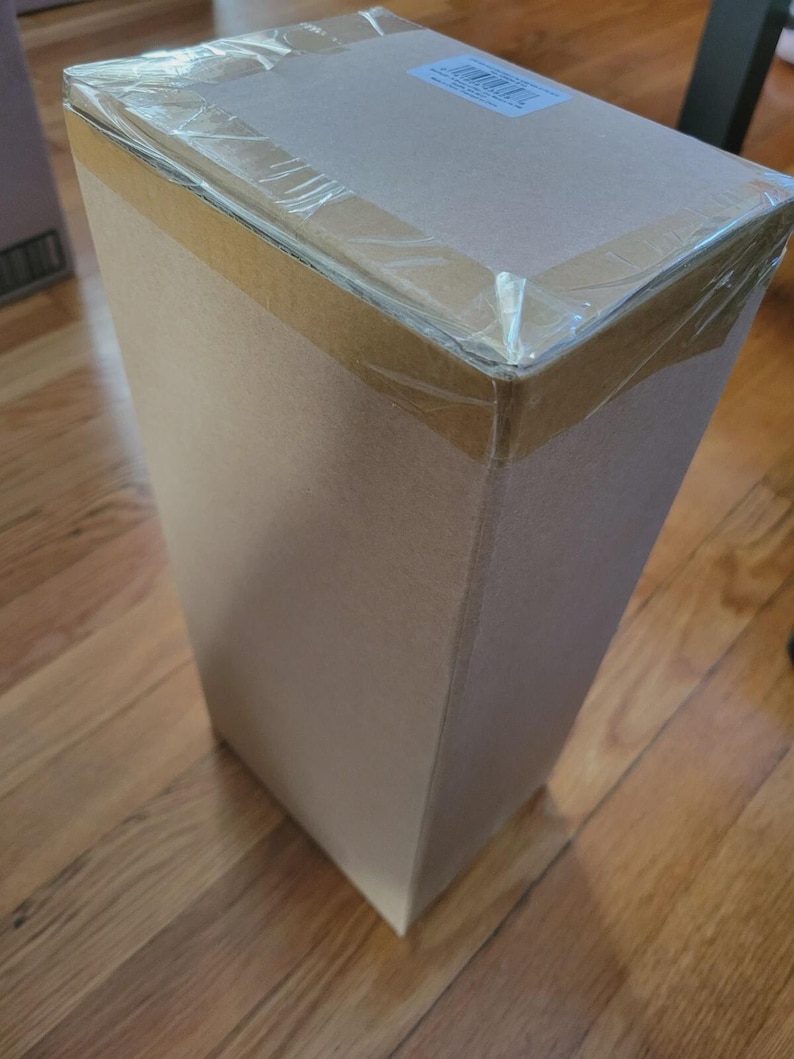 Puede incluir: Una caja de cart&oacute;n rectangular y alta, sellada con cinta adhesiva transparente. La caja es de color marr&oacute;n claro con una parte superior de color m&aacute;s claro. Una peque&ntilde;a etiqueta es visible en la parte superior. La caja est&aacute; sobre un suelo de madera.