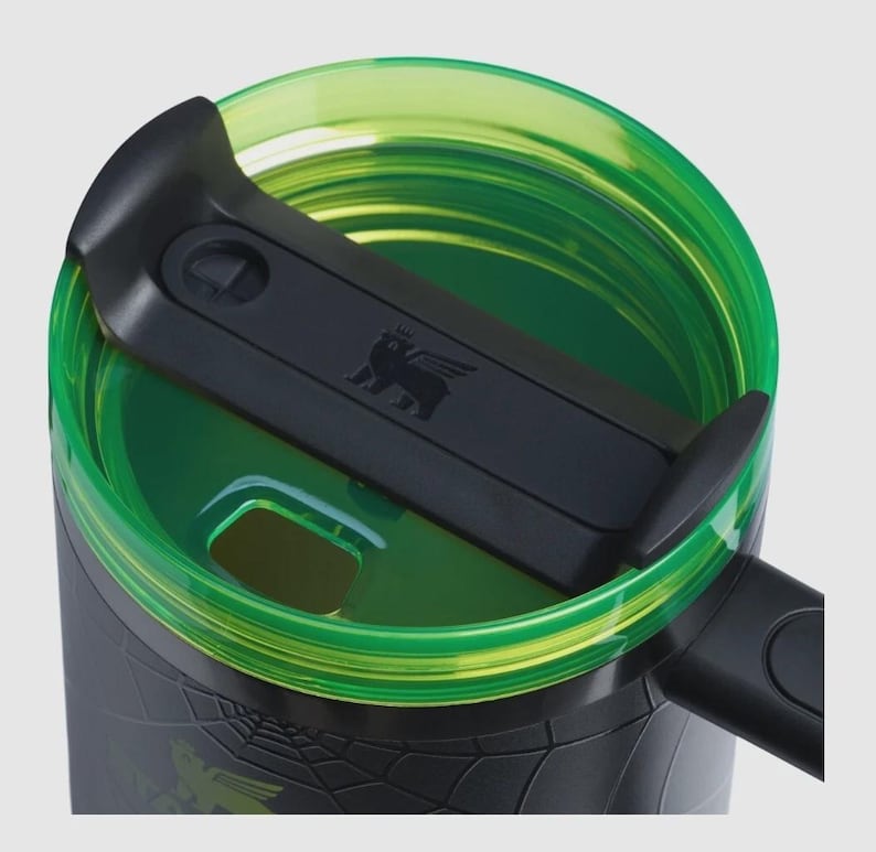 Puede incluir: Primer plano de un termo de viaje negro y verde. La tapa es negra con un peque&ntilde;o emblema y un cierre deslizante. El borde y el interior son de color verde transparente. El exterior del termo tiene un patr&oacute;n texturizado y un gr&aacute;fico verde.
