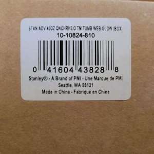 Puede incluir: Una etiqueta rectangular blanca con un c&oacute;digo de barras y texto. El texto incluye "STAN ADV 40OZ QNCHRH2.0 TM TUMB WEB GLOW (BOX)" y "Stanley&reg; - A Brand of PMI". La etiqueta est&aacute; sobre una superficie de cart&oacute;n marr&oacute;n.
