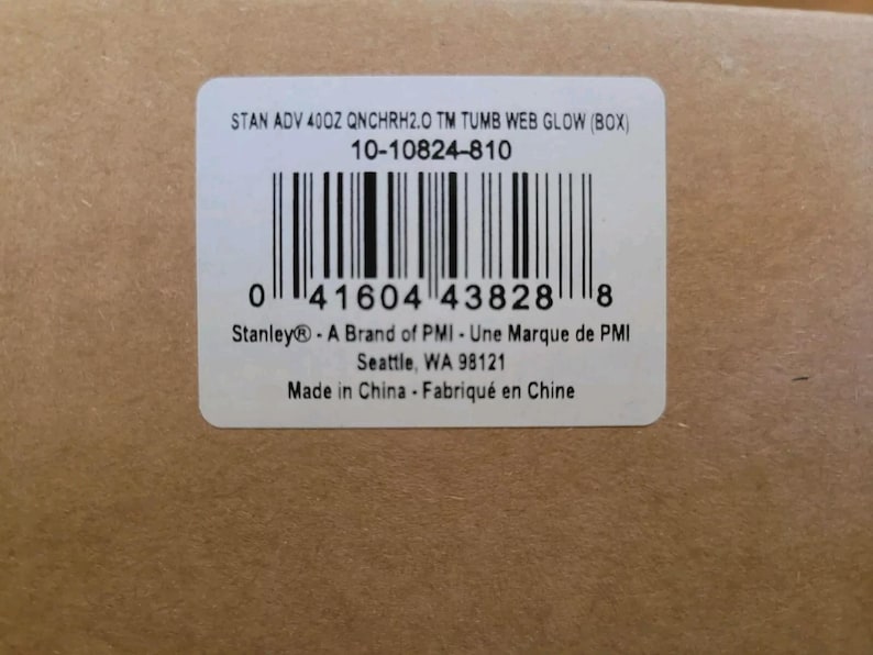 Puede incluir: Una etiqueta rectangular blanca con un c&oacute;digo de barras y texto. El texto incluye "STAN ADV 40OZ QNCHRH2.0 TM TUMB WEB GLOW (BOX)" y "Stanley&reg; - A Brand of PMI". La etiqueta est&aacute; sobre una superficie de cart&oacute;n marr&oacute;n.