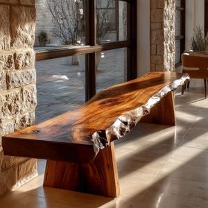 May include: A handcrafted wooden bench with a live edge. The bench is made of dark brown wood with a natural, irregular edge. It has a rectangular top and two trapezoidal legs. The bench is placed in front of a large window.