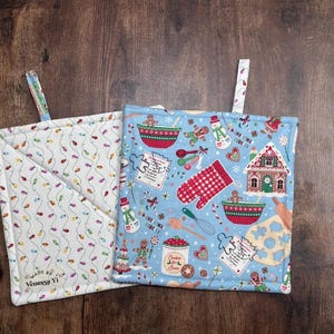May include: Two square Christmas-themed pot holders. One is white with a pattern of colorful string lights. The other is light blue with gingerbread men, a gingerbread house, and other baking-related images. Both have a loop for hanging.