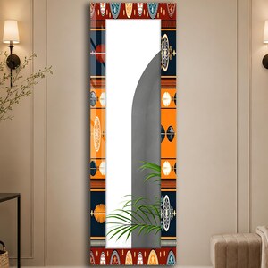 May include: A rectangular wall mirror featuring a decorative border with a repeating pattern of orange, dark blue, and white geometric shapes. The mirror surface reflects a portion of a green plant and a curved gray wall section in the room.