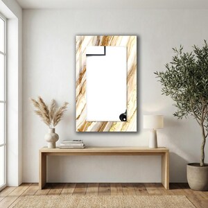 Peut inclure: Un miroir rectangulaire avec un motif marbr&eacute; dans les tons or, blanc et marron. Le miroir est dot&eacute; d'une pince noire en haut et d'un bouton noir en bas. Il est accroch&eacute; &agrave; un mur blanc.