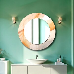 Op de afbeelding: Een ronde wandspiegel met een brede rand voorzien van een wervelend, gemarmerd patroon in tinten van zacht perzik, cr&egrave;me en lichtgrijs. De spiegel heeft een glad, reflecterend middengedeelte en is bevestigd aan een vlakke muur.