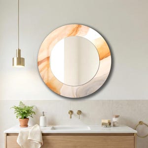 Op de afbeelding: Een ronde wandspiegel met een decoratieve rand met een abstract, vloeiend patroon in tinten warm oranje, cr&egrave;me en zachtgrijs. De spiegel heeft een glad, reflecterend midden en is gemonteerd op een effen, lichtgekleurd wandoppervlak.