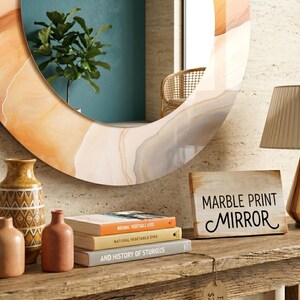 Op de afbeelding: Een ronde wandspiegel met een oranje, beige en grijs marmerpatroon frame. Onder de spiegel staan twee keramische vazen, een stapel van drie boeken en een rechthoekig houten bord met de tekst MARBLE PRINT MIRROR in het zwart gedrukt.