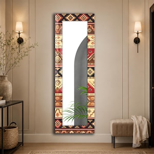 Op de afbeelding: Een rechthoekige wandspiegel met een brede lijst versierd met een herhalend geometrisch patroon in tinten rood, zwart, beige en crème. Het spiegeloppervlak toont een gedeeltelijk zicht op een grijze muur en groene palmachtige bladeren.