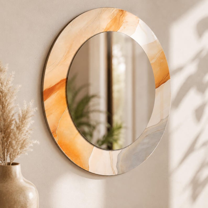 Op de afbeelding: Een ronde wandspiegel met een brede, decoratieve lijst voorzien van een abstract, vloeiend patroon in tinten van warm amber, goudoranje en zachtgrijs. Het reflecterende glazen midden biedt een helder zicht, omringd door de gladde, kleurrijke rand.