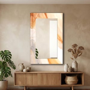 Op de afbeelding: Een rechthoekige wandspiegel met een decoratieve rand met een abstract vloeiend patroon in tinten oranje, cr&egrave;me en grijs. Het centrale reflecterende oppervlak is helder en toont een reflectie van een groen blad en een wit verticaal paneel.