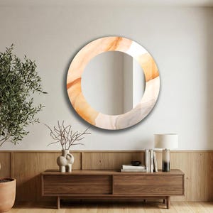 Op de afbeelding: Een ronde wandspiegel met een brede rand voorzien van een abstract, vloeiend patroon in zachte perzik-, cr&egrave;me- en lichtgrijze tinten. De spiegel heeft een glad, reflecterend midden en is bevestigd aan een effen muur boven een houten console.
