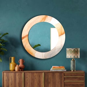Op de afbeelding: Een ronde wandspiegel met een brede lijst voorzien van een wervelend, abstract patroon in tinten cr&egrave;me, beige en zacht oranje. De spiegel reflecteert een blauwgroene muur en een deel van een wit rechthoekig frame voor een gelaagd effect.