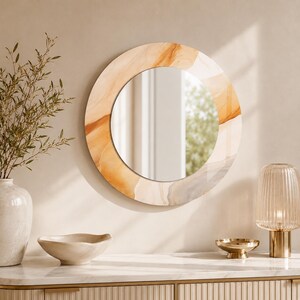 Op de afbeelding: Een ronde wandspiegel met een brede, decoratieve lijst met een wervelend, abstract patroon in tinten amber, beige en zachtgrijs. De spiegel heeft een glad, reflecterend middengedeelte en een glanzende afwerking op de buitenste rand.