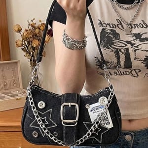 May include: Black denim shoulder bag with silver chain strap and star accents. Features a buckle closure and white stitching. Includes a small tag and silver hardware. A stylish accessory.