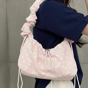 Puede incluir: Un bolso de hombro rosa con lunares blancos. El bolso tiene un cierre de cordón y un asa fruncida. El bolso está hecho de un material suave y ligero.
