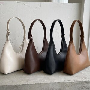 May include: Four shoulder bags in a row, each with a curved handle. The bags are in cream, brown, black, and tan. The bags are made of a smooth material and have a simple, minimalist design. The bags are sitting on a concrete surface.
