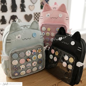 Może przedstawiać: Trzy plecaki w kształcie kota w pastelowych kolorach: jasnoniebieskim, różowym i czarnym. Każdy plecak ma kocie uszy, przeszywane detale i guzik. Przód posiada przezroczystą kieszeń wypełnioną ozdobnymi przypinkami. Małe białe skrzydła są przymocowane do boków.