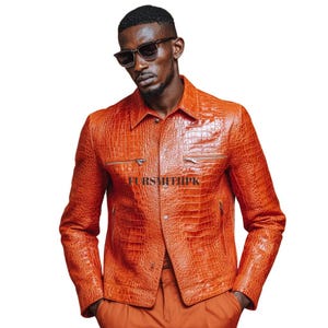 Peut inclure: Veste en cuir orange avec une texture peau de crocodile. La veste a un col, deux poches de poitrine zippées et des boutons. Le texte "FURSMITHPK" est imprimé sur la veste.
