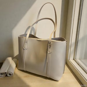 Pode incluir: Uma bolsa tote cinza claro com duas alças e formato retangular. A bolsa tem uma textura lisa e detalhes de costura verticais. Um pano enrolado é visível no lado esquerdo.