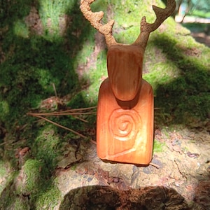 May include: A hand-carved wooden figurine of a deer with a smooth, light brown body featuring a carved spiral design on the chest. The deer has textured, branch-like antlers extending upwards from its head, showcasing natural wood grain details.