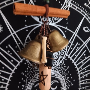 Könnte beinhalten: Ein handgefertigter Talisman mit Zimtstange, Messingglocken, einem Knochen und einem schwarzen Stein. Der Talisman hängt an einer braunen Schnur mit einer grünen Perle. Am unteren Ende befindet sich ein Holzylinder mit einem Pentagramm.