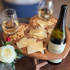 May include: A wooden heart-shaped serving tray with compartments for cheese, crackers, and nuts. A bottle of wine and two glasses of white wine are on the tray. A small vase with a white rose and a red rose is on the side.