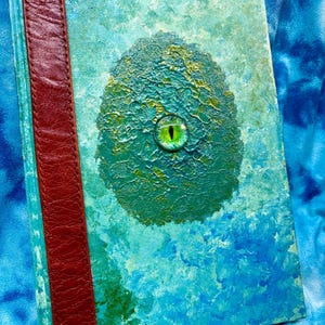 Peut inclure: Un journal fait à la main avec une couverture texturée bleu sarcelle et bleue, avec un ovale vert texturé en relief avec un œil vert. Une tranche en cuir bordeaux longe le côté gauche.