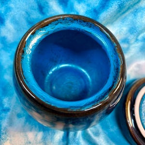 May include: A ceramic jar with a lid, featuring a vibrant blue interior and a glossy, dark brown rim. The jar's exterior has a textured appearance, and the lid is partially visible, showcasing a matching design.