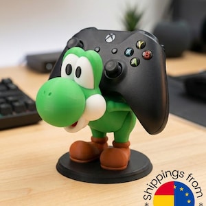 Puede incluir: Una figura verde y blanca de Yoshi sosteniendo un mando de Xbox negro. La figura tiene botas marrones y una base circular negra. El mando tiene botones de colores y descansa sobre la espalda de la figura.