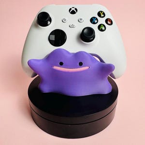 Op de afbeelding: Een witte Xbox-controller met zwarte knoppen en gekleurde actieknoppen staat boven een paarse Ditto-figuur. De Ditto-figuur staat op een zwarte basis, alles tegen een roze achtergrond. De controller heeft een zwart kruis en twee zwarte joysticks.
