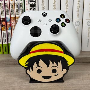 Può includere: Un controller wireless bianco per videogiochi appoggiato su un supporto in plastica nera a forma di testa di personaggio dei cartoni animati. Il personaggio ha capelli neri, un cappello di paglia giallo con fascia rossa e una cicatrice.