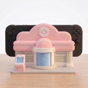 Puede incluir: Un soporte decorativo rosa y blanco con forma de edificio, dise&ntilde;ado para sostener una consola Nintendo Switch negra. El soporte presenta una peque&ntilde;a m&aacute;quina recreativa rosa a juego. El edificio tiene una puerta azul claro y un logotipo de Pokemon blanco.