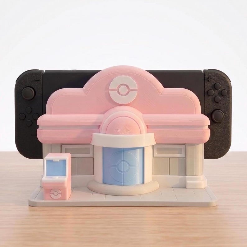 Puede incluir: Un soporte decorativo rosa y blanco con forma de edificio, dise&ntilde;ado para sostener una consola Nintendo Switch negra. El soporte presenta una peque&ntilde;a m&aacute;quina recreativa rosa a juego. El edificio tiene una puerta azul claro y un logotipo de Pokemon blanco.