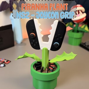 May include: A green plastic plant pot base with a central stem and two leaves, featuring a white Piranha Plant-shaped Joy-Con grip holding two black game controllers. The pot includes slots for game cartridges. Text reads: PIRANHA PLANT NS2 - JOYCON GRIP.