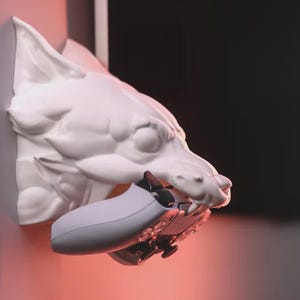 Könnte beinhalten: Eine weiße Wolfskopfskulptur, die einen grauen und schwarzen Videospiel-Controller im Maul hält. Die Skulptur ist an einer Wand befestigt. Der Hintergrund ist rosa und schwarz.